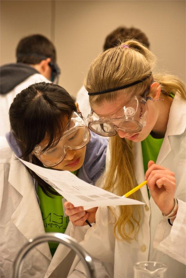 Two people wearing goggles peer at a piece of paper held by one person. That person also is holding a pencil.