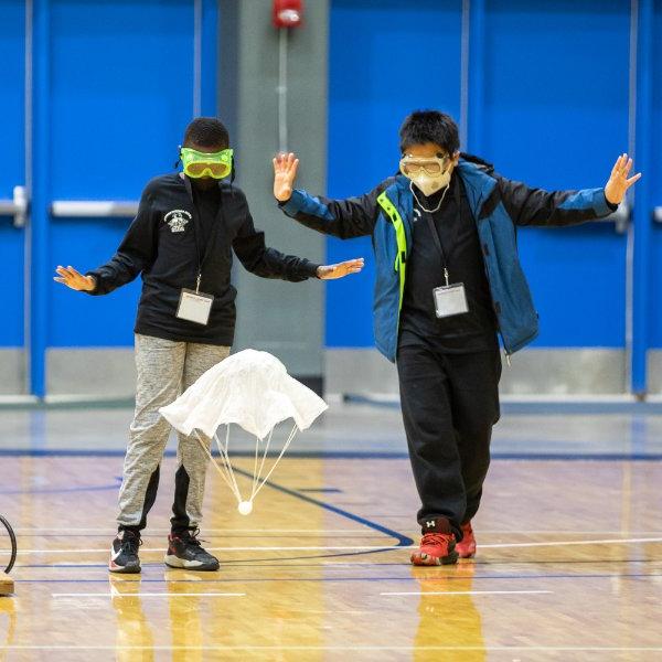 Two students, wearing goggles and masks, lift their hands in the air as they watch an object float upward.